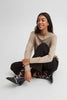 A woman sits cross-legged on a light gray studio floor, wearing a beige long-sleeve top, black pants, and black western-style boots with white stitching.